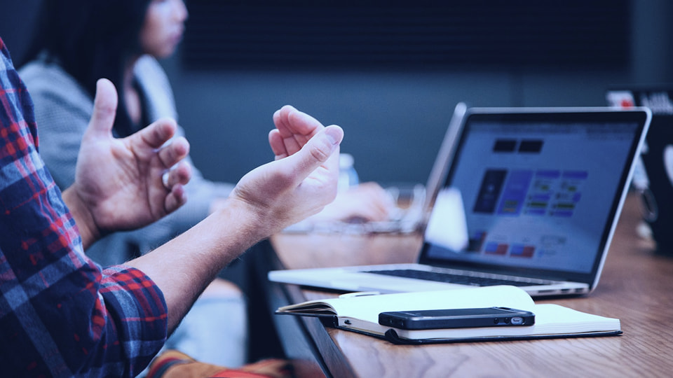 A person’s hands gesturing over a wooden conference table beside an open laptop, notebook, and smartphone, with a blurred colleague in the background.