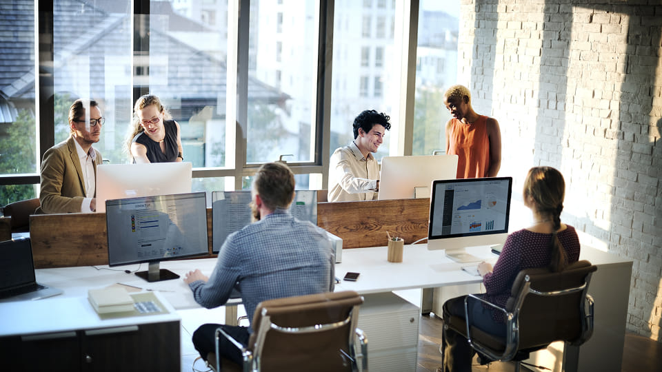 A modern open-plan office with several employees collaborating at workstations by large windows.